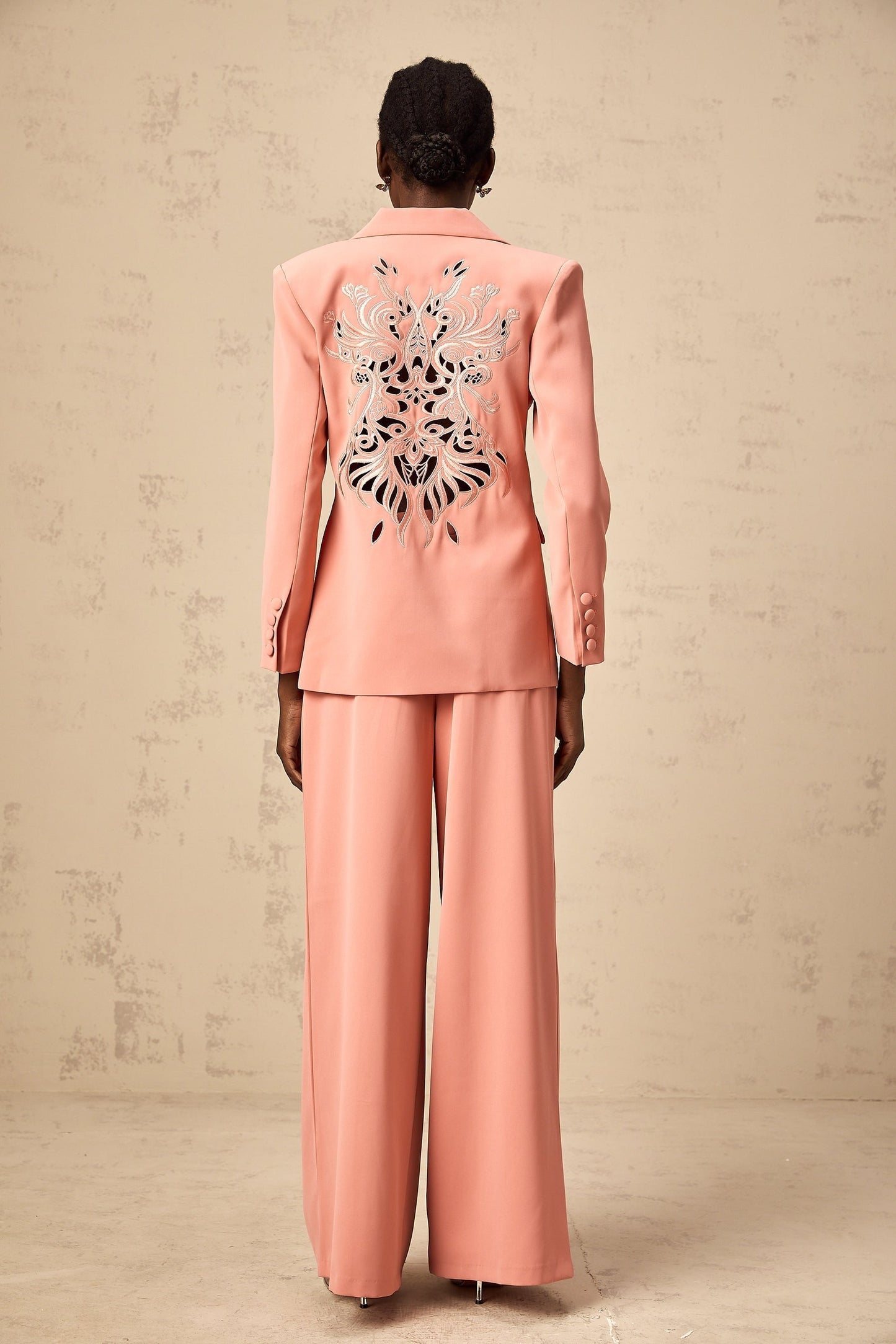 A pink blazer and trouser set with embroidery details and cutout designs The blazer features a tailored silhouette with decorative cutouts while the matching trousers have a sleek fit Both pieces are adorned with intricate pink embroidery creating an elegant and coordinated outfit