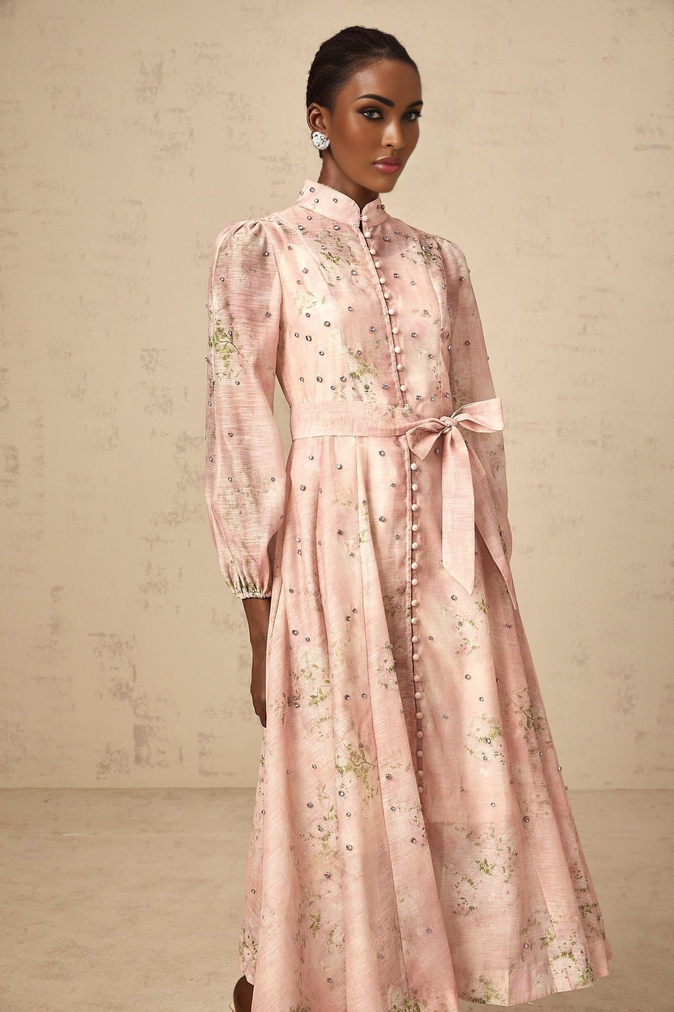 A woman wearing a pink midi dress with floral print and embellishments standing against a plain background