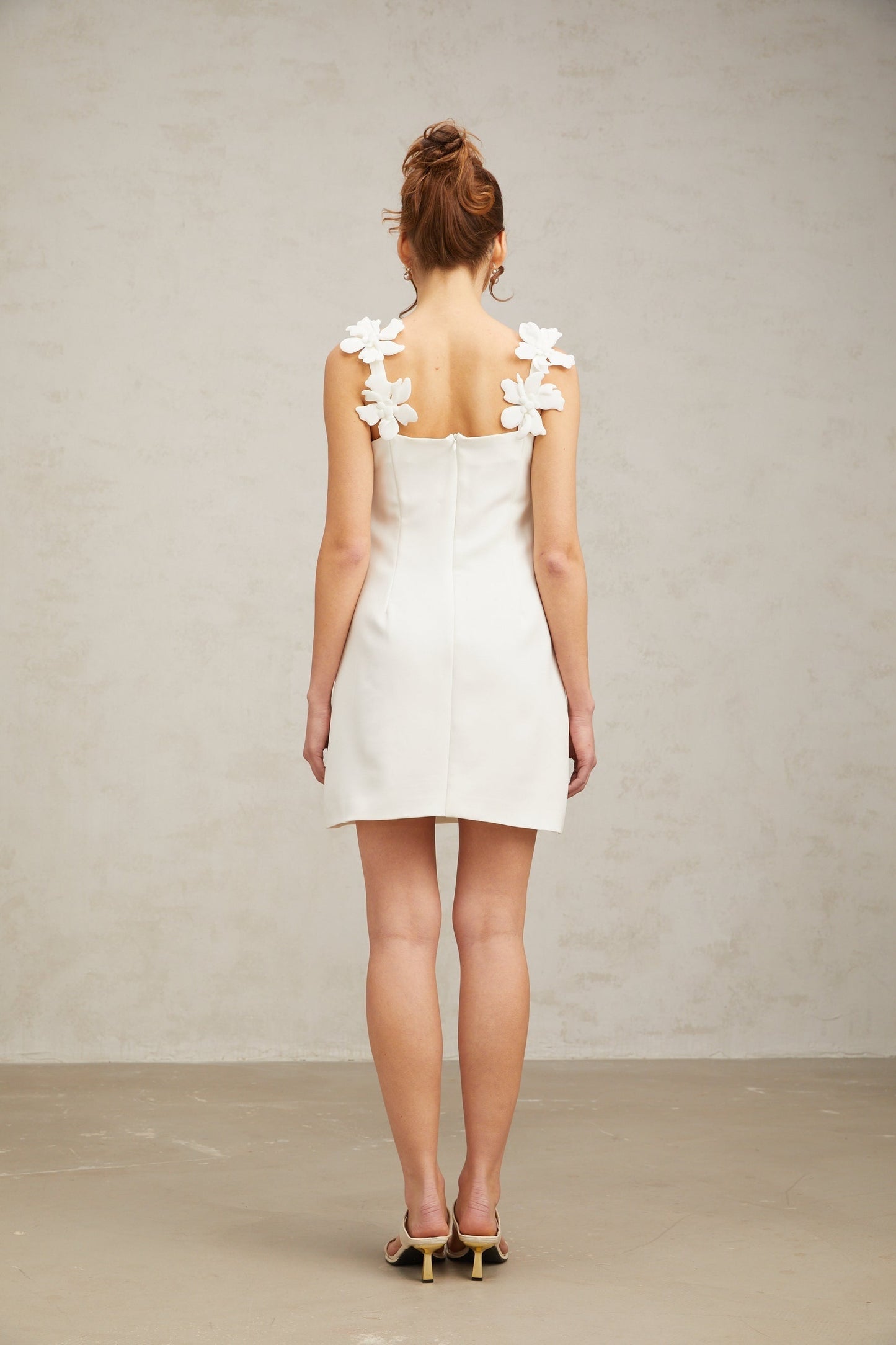 Young woman wearing a short white dress with floral embroidery standing in front of a plain background