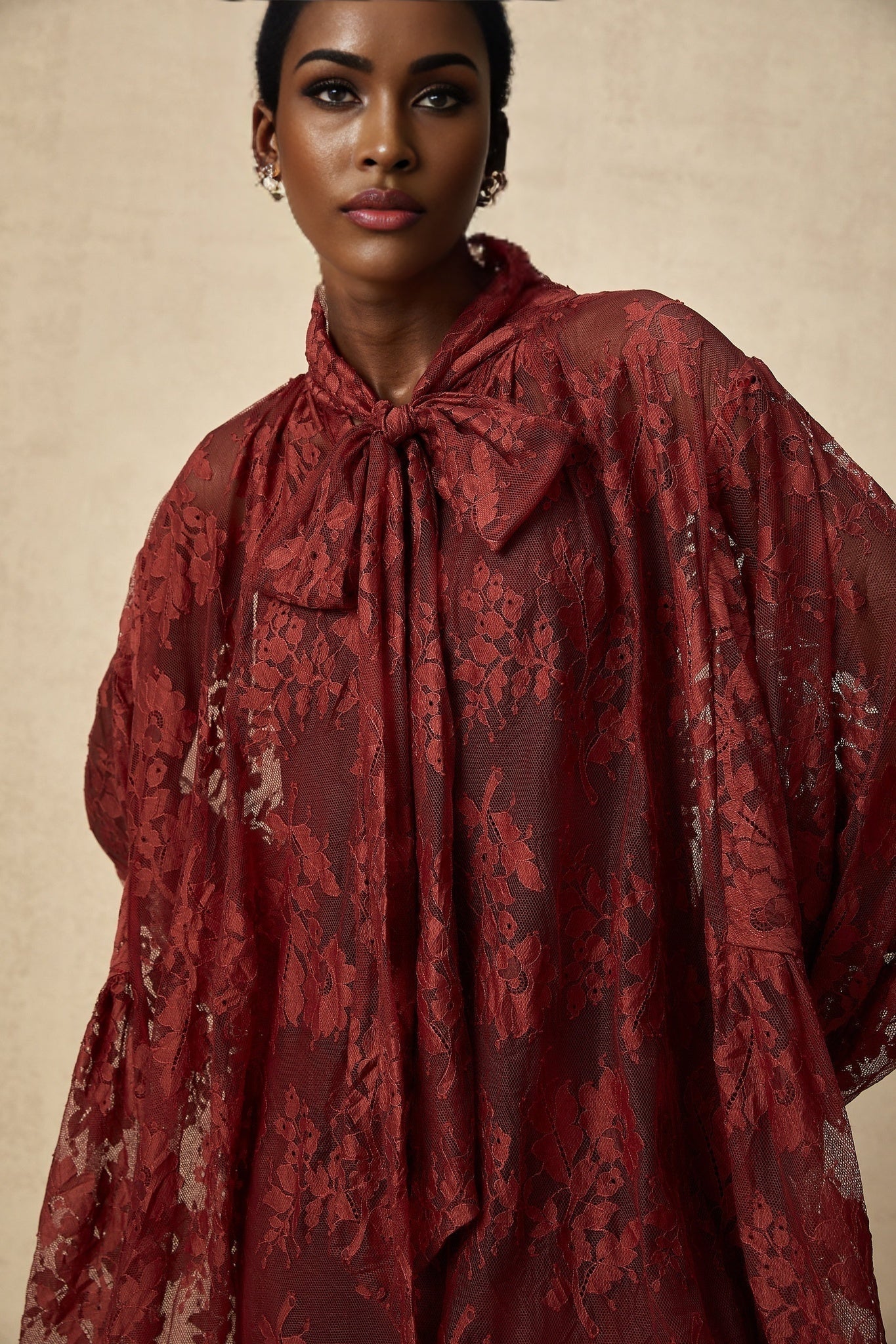 A woman wearing a long flowing red dress made of chantilly lace with an oversized fit standing against a plain background