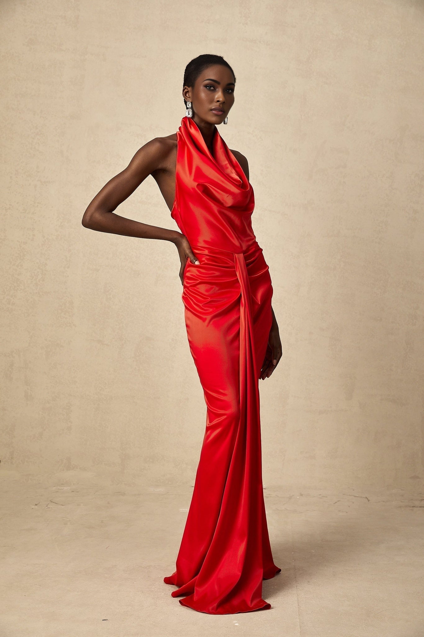 A woman wearing a long red satin dress with a halter neckline and draped fabric detail standing against a plain background