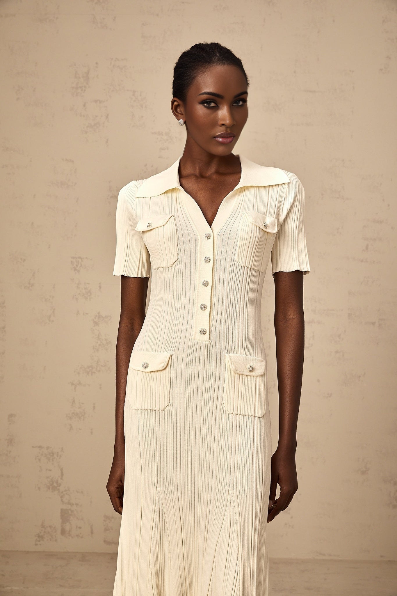 Woman wearing a beige ribbedknit midi dress with long sleeves and a slim fit silhouette standing against a plain background