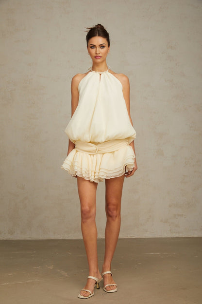 Young woman wearing a white halterneck mini dress adorned with faux pearls standing against a plain background