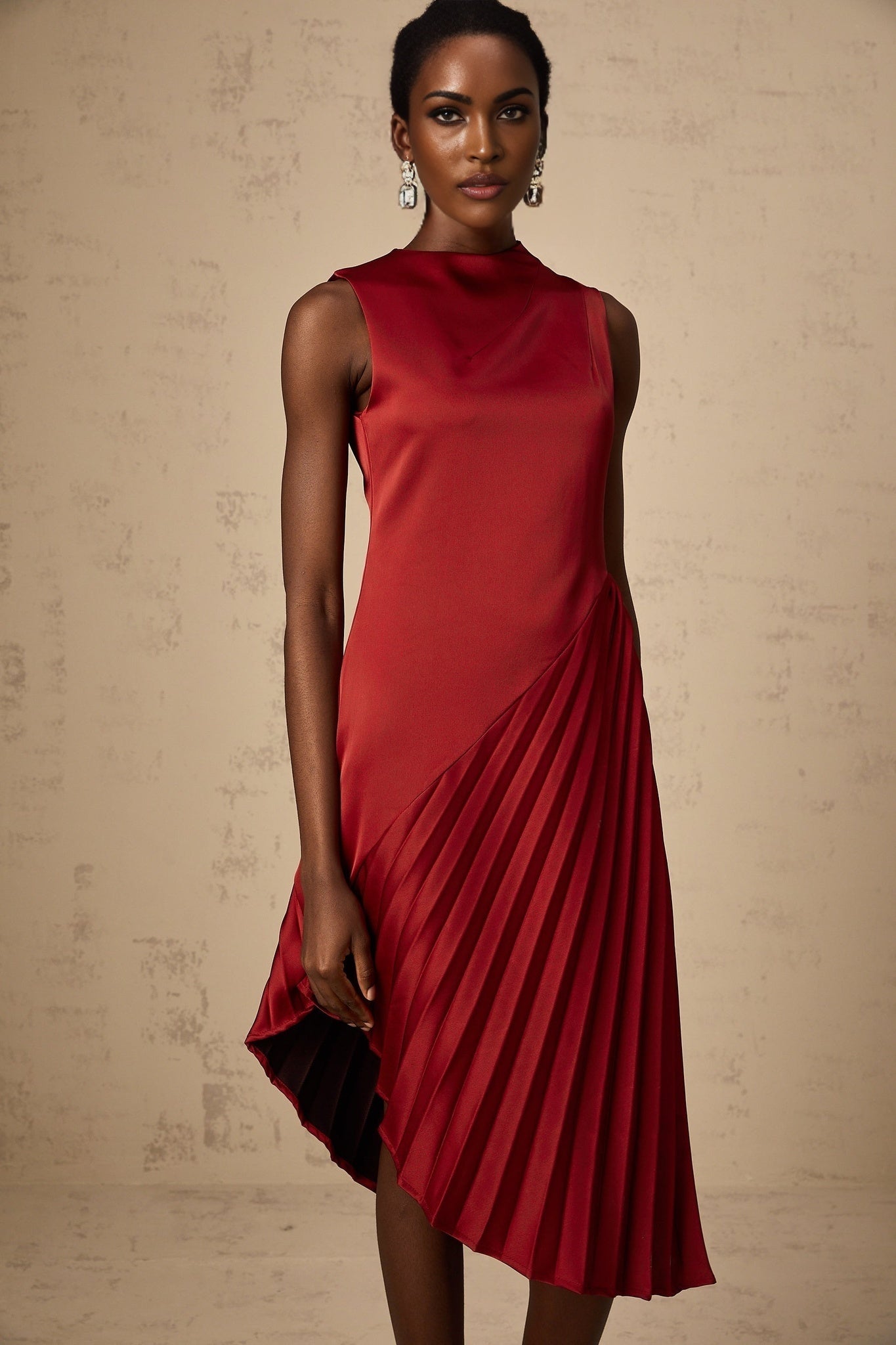 A woman wearing a wine red midi dress with an asymmetric neckline and pleated skirt standing against a plain background