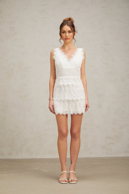 Young woman wearing a white lace mini dress with tiered skirt and short sleeves standing against a plain background