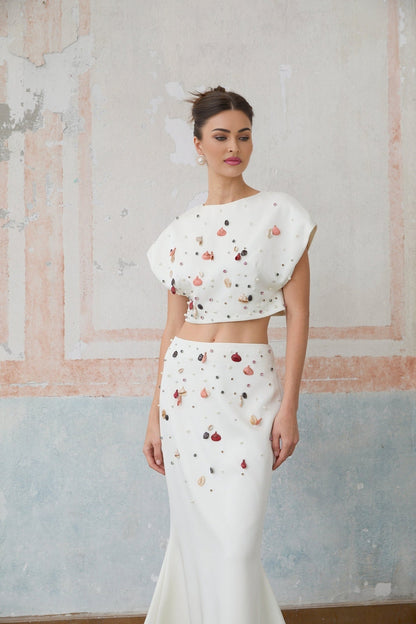 A white twopiece outfit featuring a top and skirt adorned with decorative shell embellishments both pieces matching to create a coordinated set
