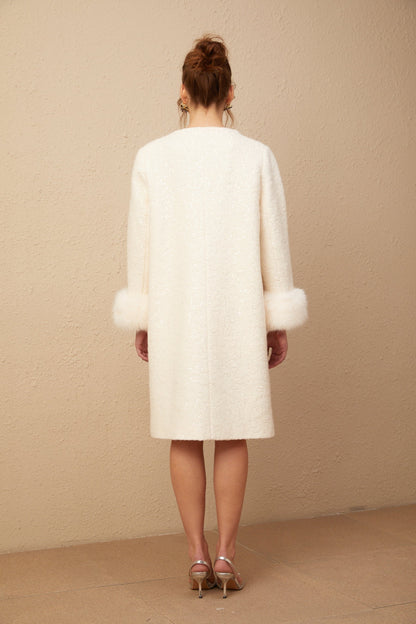Woman wearing a white collarless textured fur coat standing against a plain background