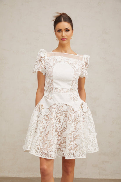 Young woman wearing a white mini dress with floral lace pattern and puff sleeves standing against a plain background