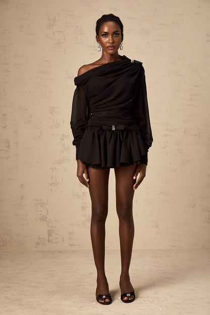 Young woman wearing black asymmetric tulle blouse and matching skirt set standing against plain background
