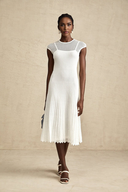 A white midi dress with scalloped hem and semi sheer fabric worn by a model standing in a neutral background