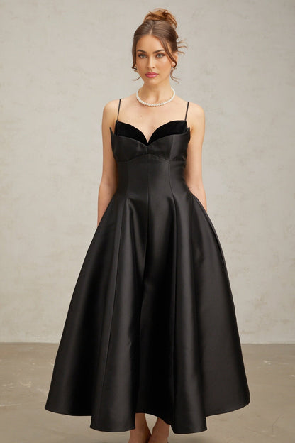 Woman wearing a black pleated midi dress with flared skirt and fitted waist standing against plain background
