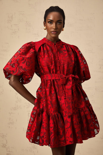 A woman wearing a short red jacquard dress with a semi sheer design standing in a relaxed pose