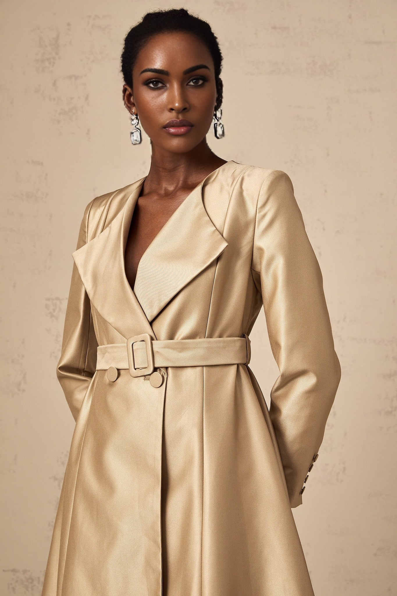 Woman wearing long khaki coat with belt at waist standing in urban setting