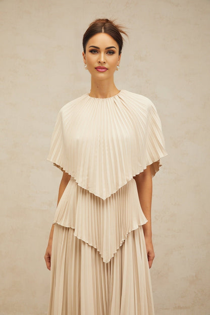 A woman wearing a flowing neutralcolored midi dress with cape detailing and pleated skirt standing in a relaxed pose