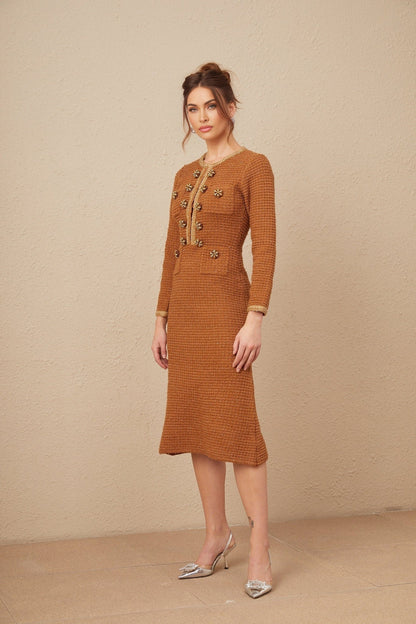 A woman wearing a brown tweed dress with braided edges standing in a neutral background