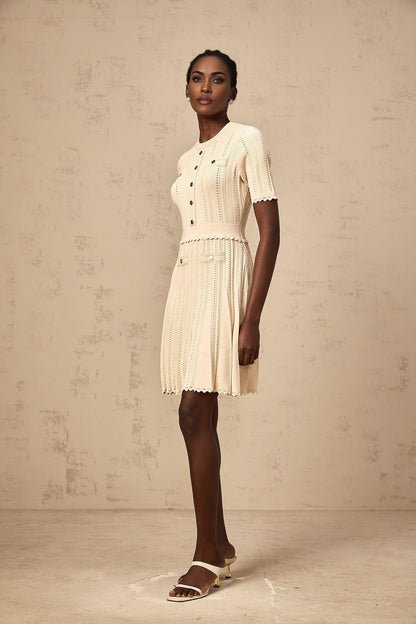 Woman wearing a lightcolored knit mini dress with pleated skirt and delicate pointelle trim detail standing in a neutral setting