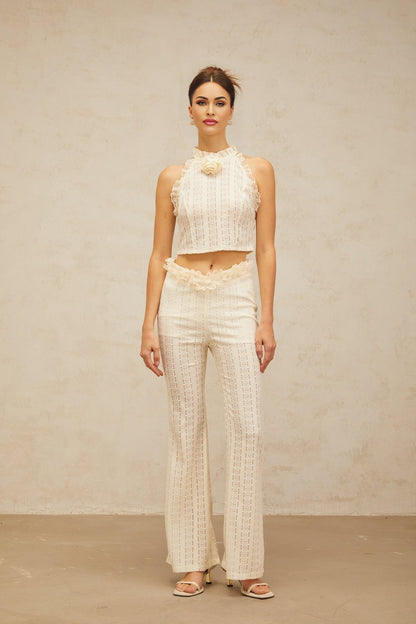 Woman wearing cream white jacquard matching set with ruffled lace trim top and trousers standing against plain background