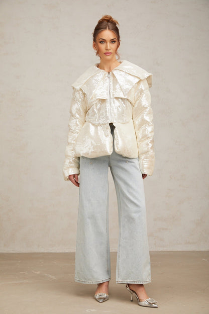 A woman wearing a white puffy down coat covered in sparkling sequins standing against a plain background