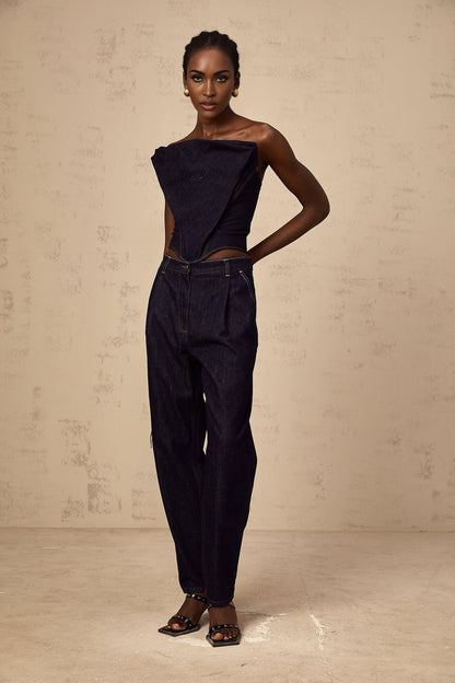 Woman wearing blue high rise jeans with pleated details on the front standing against a plain background