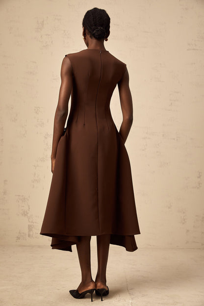 A woman wearing a brown asymmetric midi dress with one shoulder design standing against a plain background