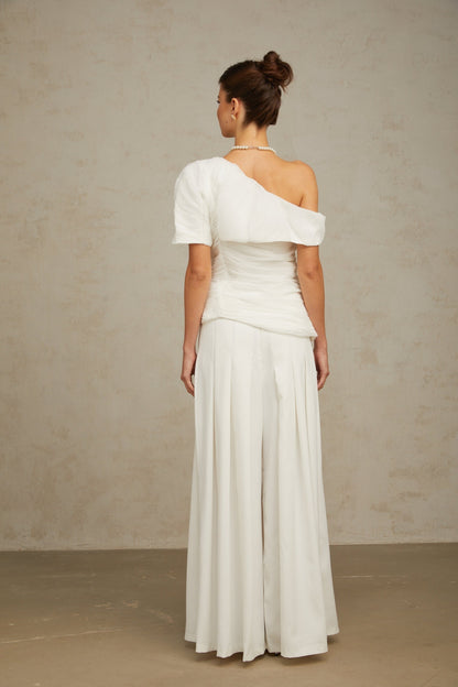 Woman wearing white matching set with creased top and trousers standing against plain background