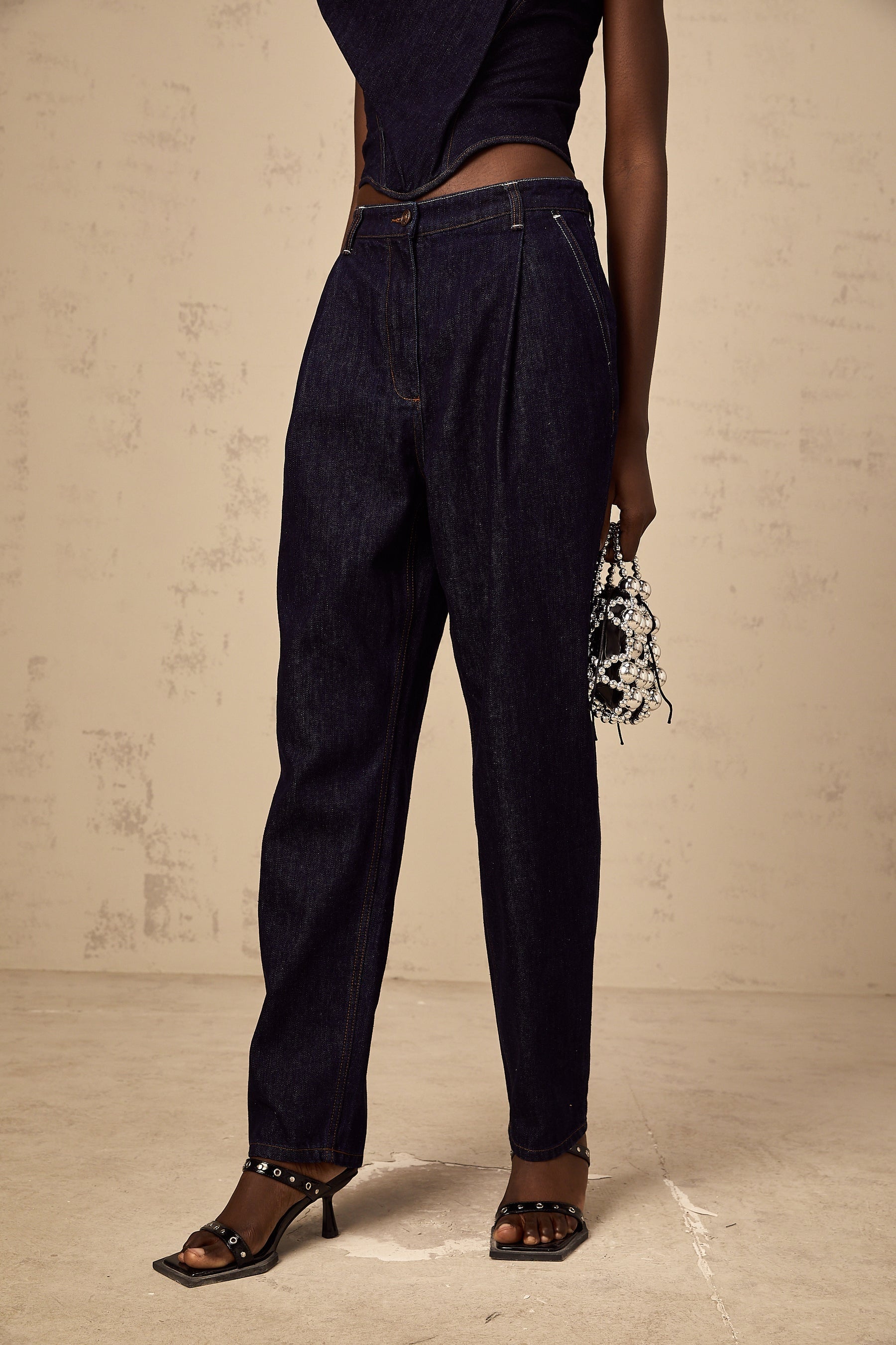 Blue high rise jeans with pleated details on the front worn by a model standing against a plain background
