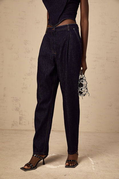 Blue high rise jeans with pleated details on the front worn by a model standing against a plain background