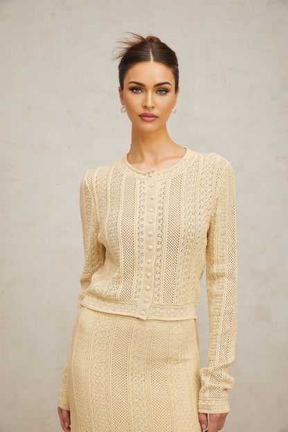 A woman wearing a matching cream colored pointelle knit cardigan and skirt set The outfit has delicate lacy patterns throughout