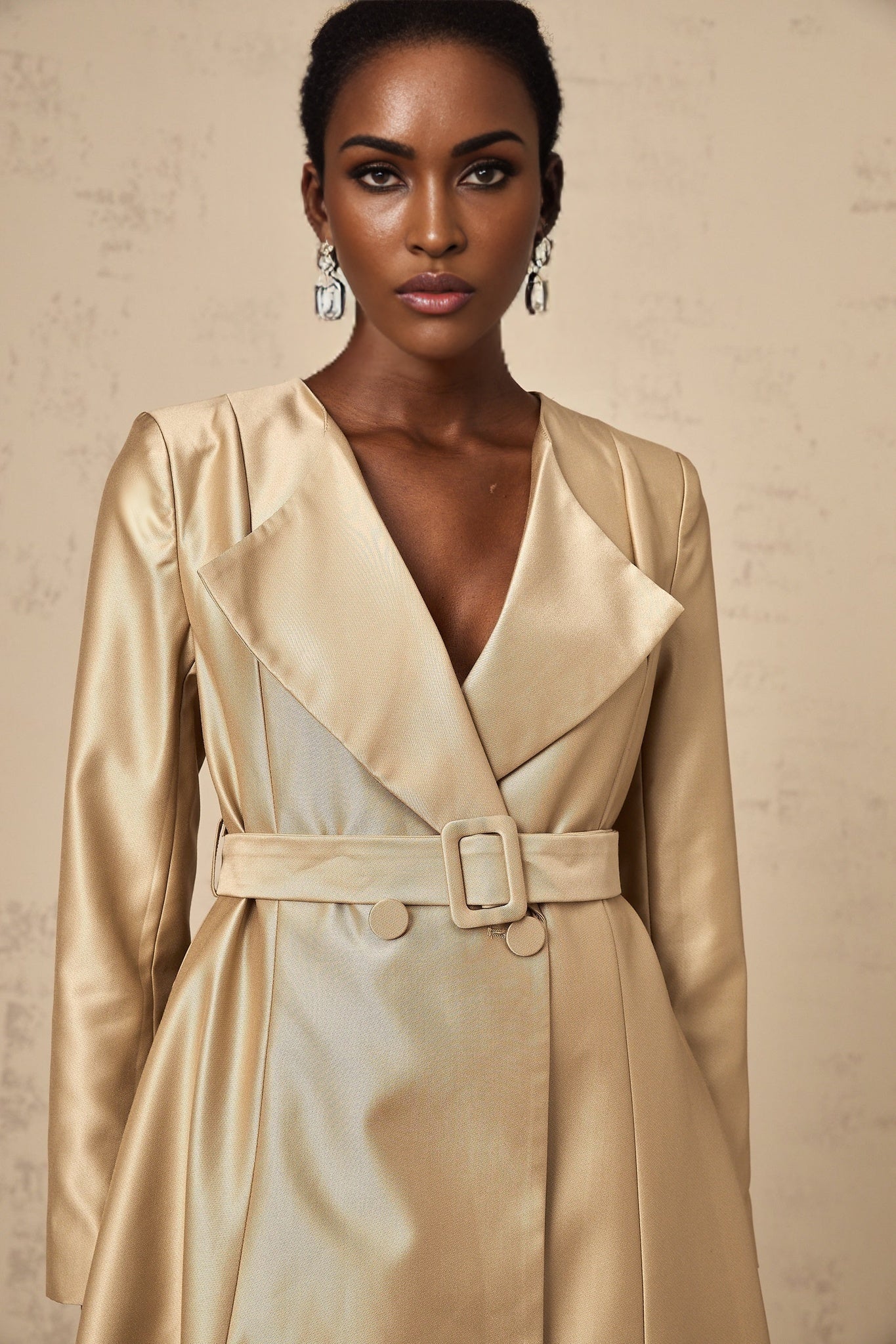 Woman wearing long khaki coat with belt at waist standing in urban setting