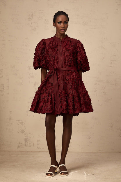 A woman wearing a short winered dress with three dimensional petal like puffs covering the fabric creating texture and volume