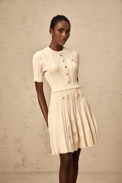 Woman wearing a beige mini dress with pleated skirt and delicate pointelle trim detail standing in a neutral setting