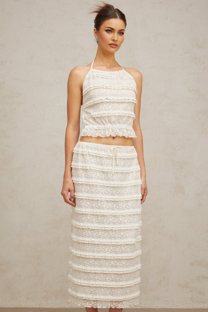 A woman wearing an off white floral lace matching set consisting of a ruffled top and skirt The outfit has delicate lace details and a feminine ruffled design