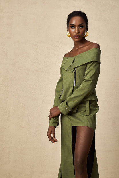 A woman wearing a green asymmetric jacket paired with a matching skirt standing against a plain background