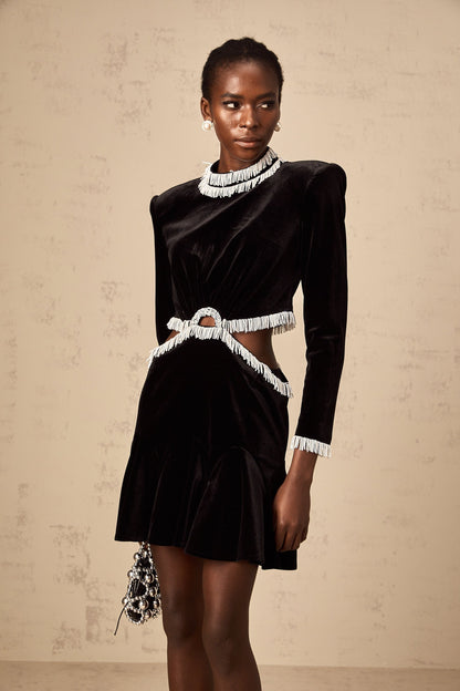 A black velvet mini dress with cut out details and fringe trim worn by a model against a plain background