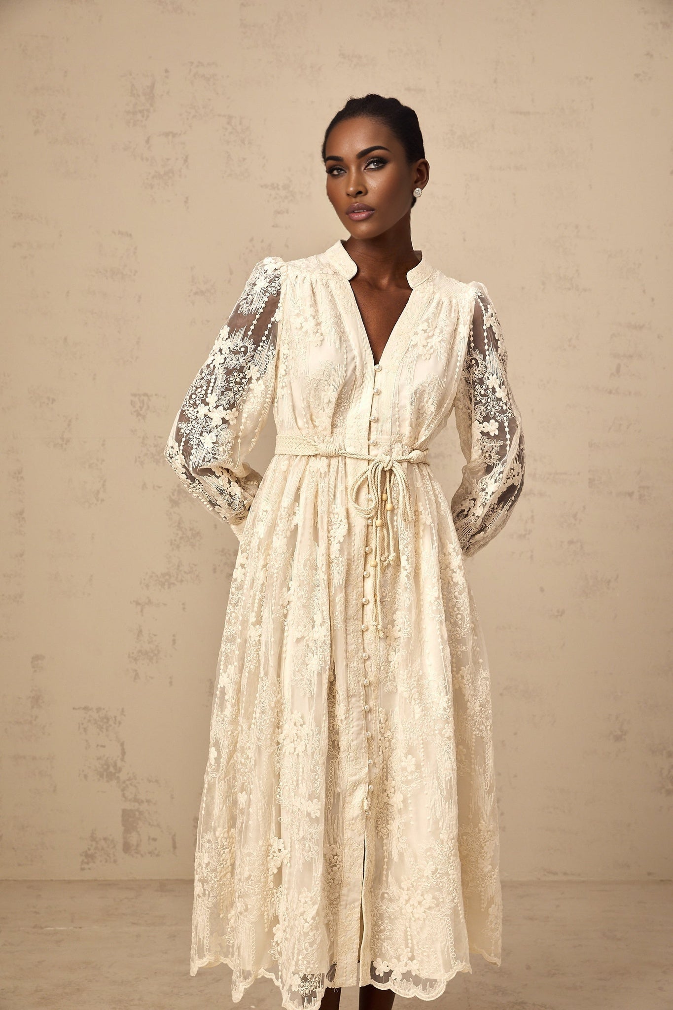 A woman wearing a neutralcolored midi dress with delicate lace embroidery details standing against a plain background