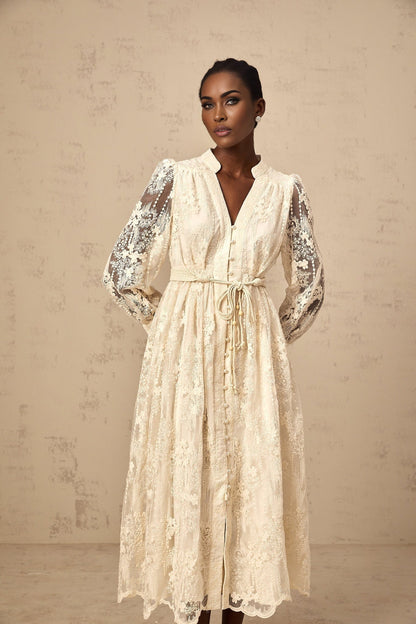 A woman wearing a neutralcolored midi dress with delicate lace embroidery details standing against a plain background