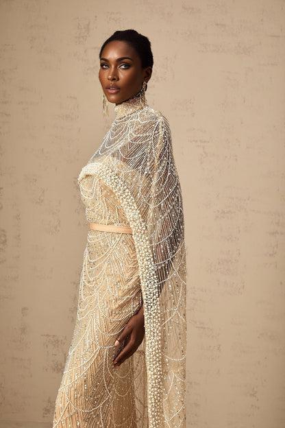 A woman wearing a champagne colored gown with a matching beaded cape The dress is floor length with a fitted silhouette while the cape drapes elegantly over the shoulders Both pieces feature intricate beadwork that sparkles in the light