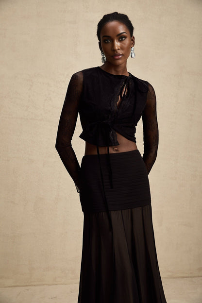 A black cropped top with sheer lace trim and see through details worn by a model against a plain background