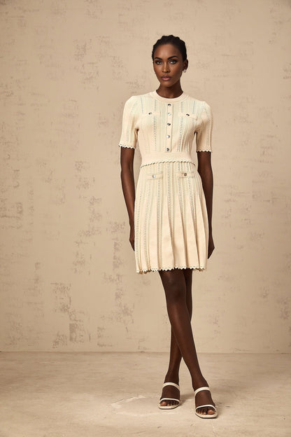 A woman wearing a short neutral colored knit dress with pleated skirt and delicate pointelle trim detailing