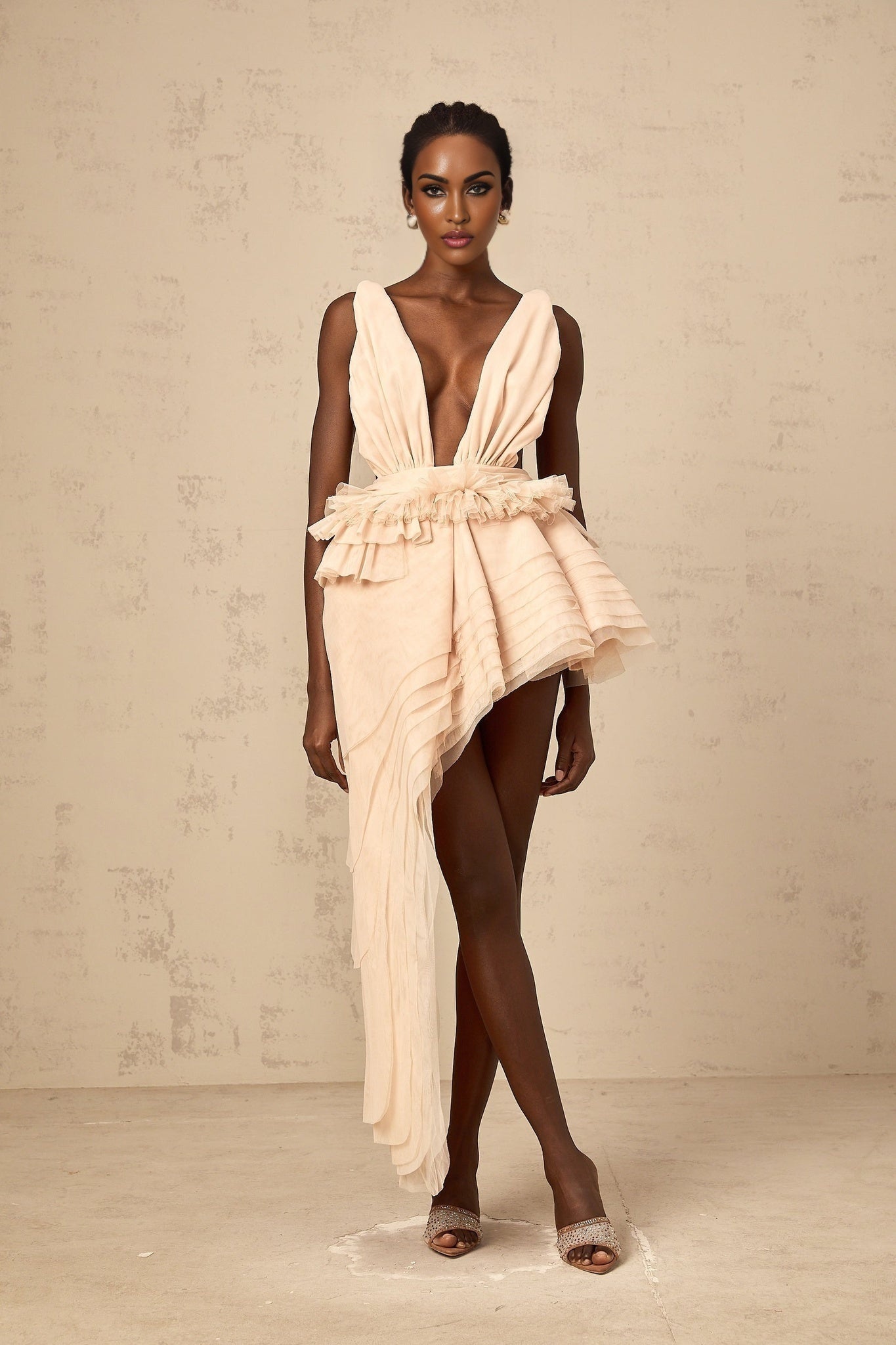 A woman wearing a lightcolored asymmetric mini dress with ribbon design details standing against a plain background
