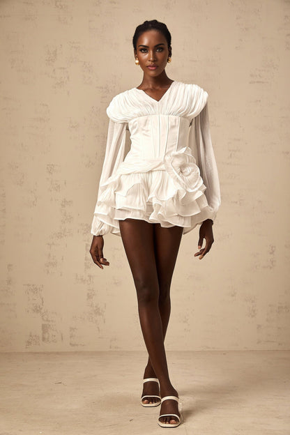 Young woman wearing a white fully pleated mini dress with cinched waist detail standing against a plain background