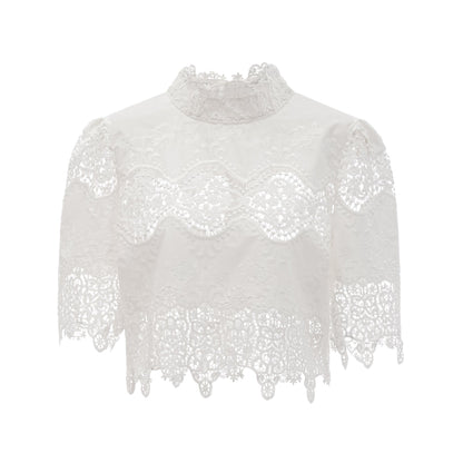 Woman wearing white lace trim cropped top and matching trousers with cutout details standing against plain background