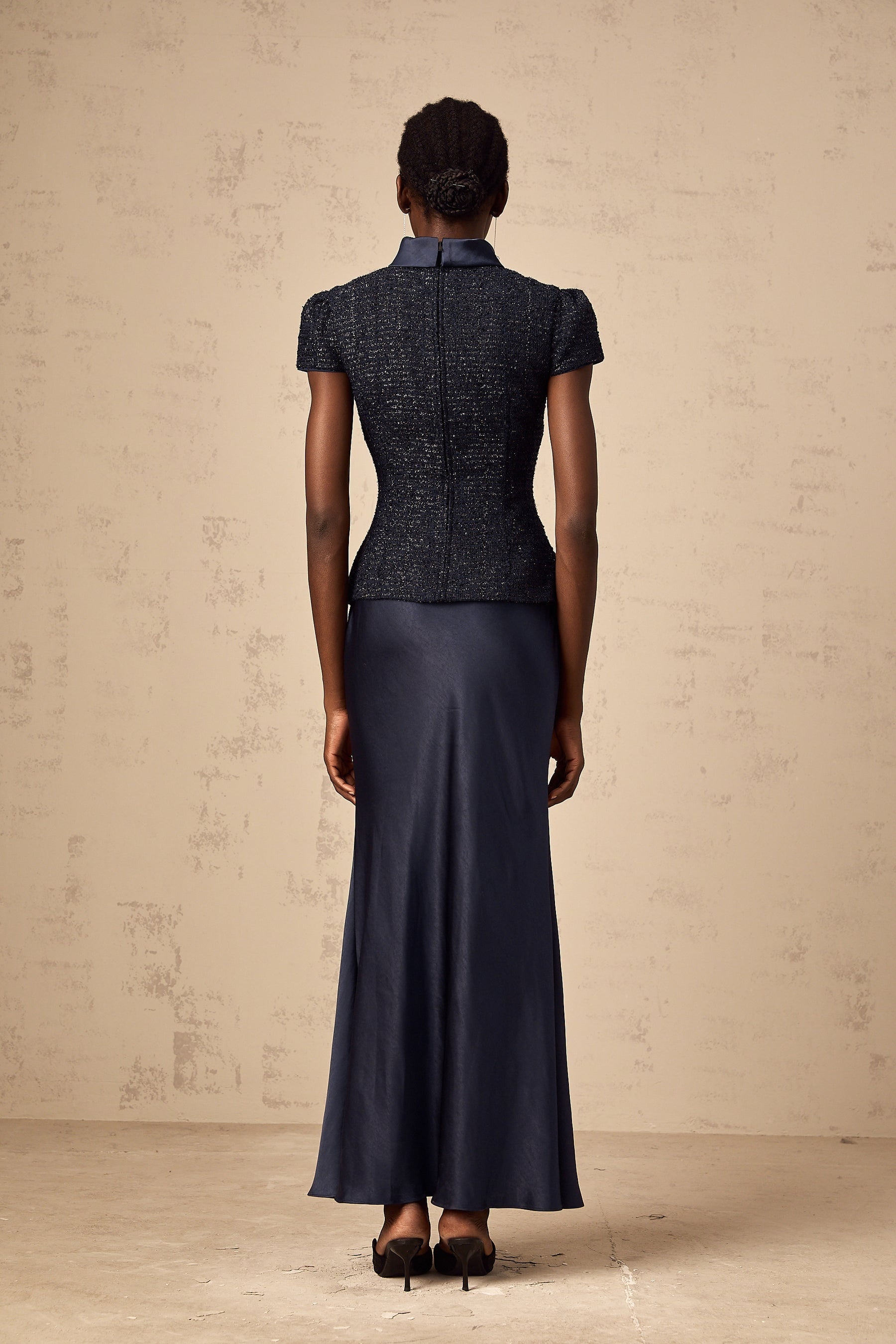 A woman wearing a long blue maxi dress with a fitted peplum waist and paneled design standing against a plain background