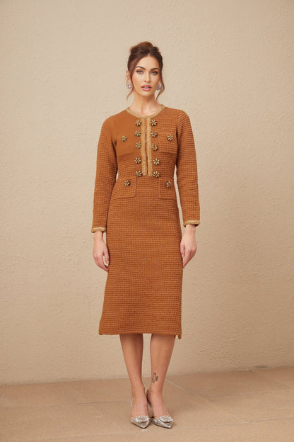 A woman wearing a brown tweed dress with braided edges and a fitted silhouette standing against a plain background