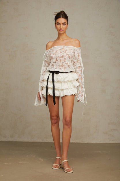 A woman wearing a white floral lace matching set consisting of a cropped top and skirt standing against a plain background