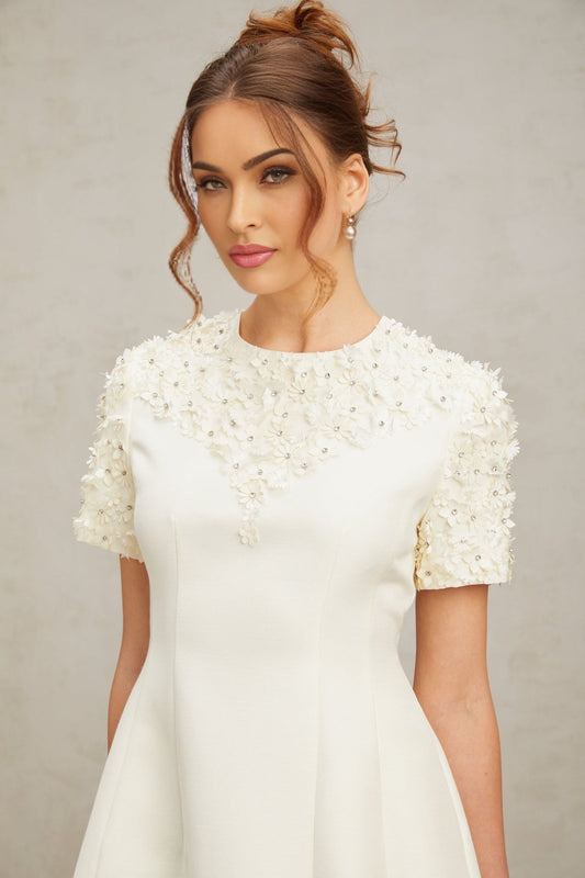 A short white mini dress with colorful floral appliqués scattered across the fabric featuring a fitted bodice and flared skirt