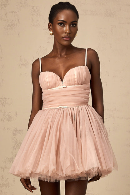 Young woman wearing a sleeveless pleated tulle mini dress in a light color standing against a plain background