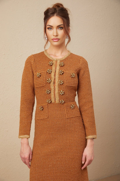 A woman wearing a brown tweed dress with braided edges standing against a plain background