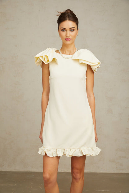 Young woman wearing a short white mini dress with ruffled details and a large bow accent standing against a plain background