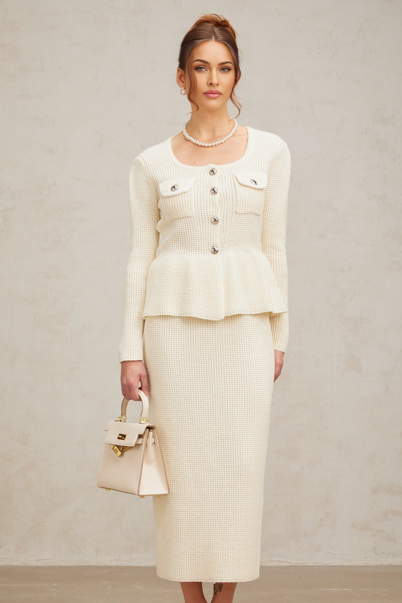 A woman wearing a white knitted matching set consisting of a sleeveless top and a knee length skirt standing against a plain background